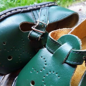 Pode incluir: Um par de sapatos de couro verde com fivelas e uma tira. Os sapatos t&ecirc;m um detalhe de costura branca ao redor da borda e um padr&atilde;o de pequenos furos.