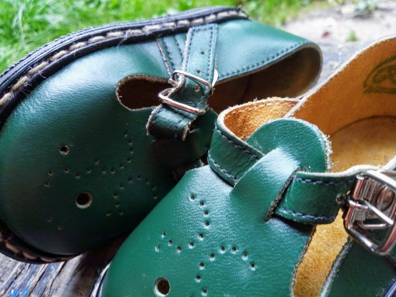 Pode incluir: Um par de sapatos de couro verde com fivelas e uma tira. Os sapatos t&ecirc;m um detalhe de costura branca ao redor da borda e um padr&atilde;o de pequenos furos.