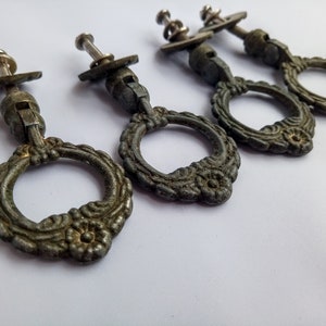 Könnte beinhalten: Vier antike Schubladengriffe mit verzierten Metalldesigns. Die Griffe sind dunkelgrau und haben eine runde Ringform mit einem dekorativen Blumenmuster.