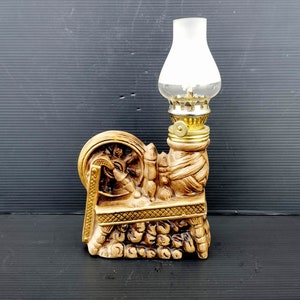 May include: A ceramic oil lamp shaped like a spinning wheel with a clear glass chimney. The lamp is painted in shades of brown and gold.