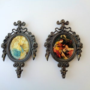 May include: Two antique oval picture frames with ornate bronze-colored metalwork. Each frame holds a small, colorful print of classical art. One depicts a woman in prayer, the other a couple.