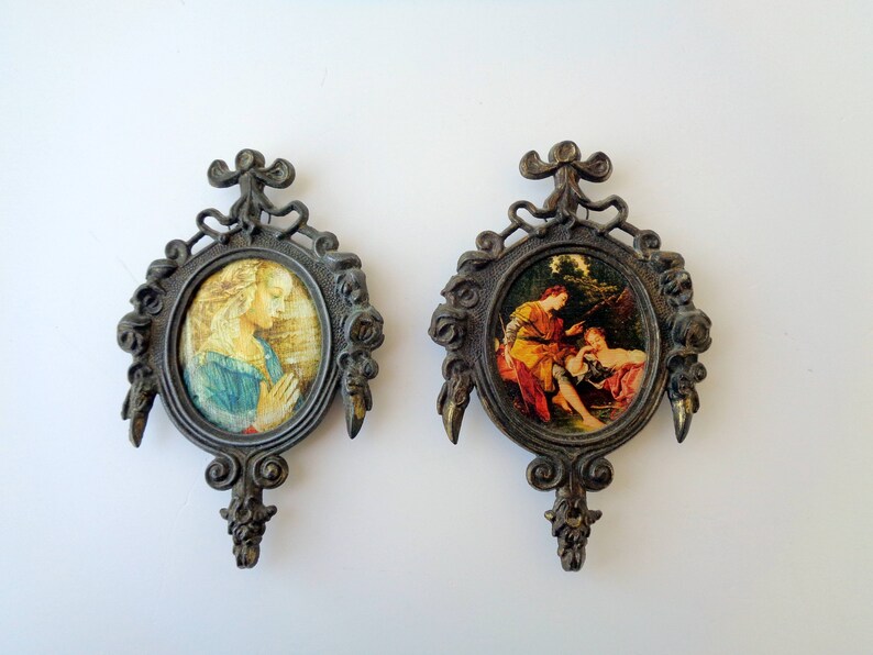 May include: Two antique oval picture frames with ornate bronze-colored metalwork. Each frame holds a small, colorful print of classical art. One depicts a woman in prayer, the other a couple.