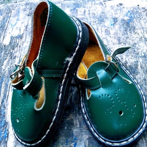 Pode incluir: Um par de sapatos infantis de couro verde escuro com costuras brancas e fecho de fivela. Os sapatos t&ecirc;m uma ponta redonda e uma sola grossa.