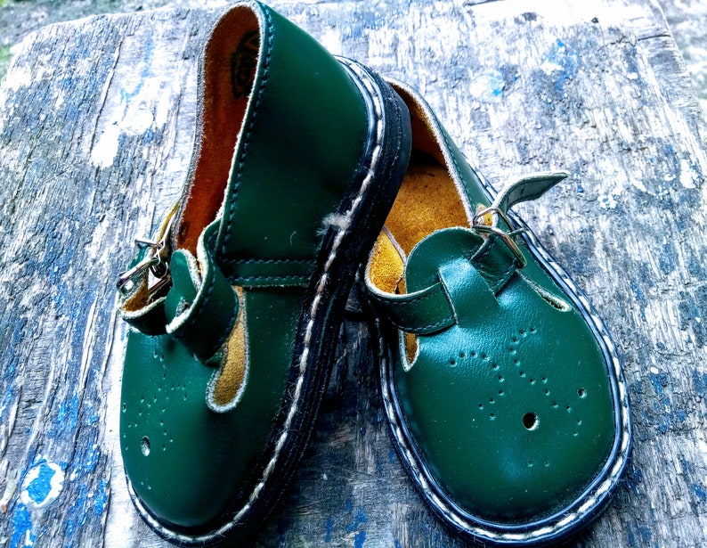 Pode incluir: Um par de sapatos infantis de couro verde escuro com costuras brancas e fecho de fivela. Os sapatos t&ecirc;m uma ponta redonda e uma sola grossa.