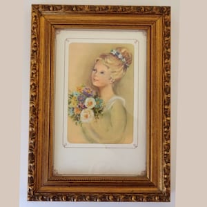 May include: A framed print of a young girl with blonde hair holding a bouquet of flowers. The girl is wearing a green dress and has a sweet expression on her face. The frame is gold and has a decorative design.
