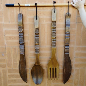 Könnte beinhalten: Vier hölzerne Küchenutensilien hängen an einer Wand. Die Utensilien sind ein Messer, ein Löffel, eine Gabel und ein weiteres Messer. Die Utensilien sind alle mit komplizierten Mustern geschnitzt.