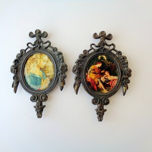 May include: Two antique oval wall plaques with ornate, dark metal frames. Each plaque features a classical painting. One depicts a woman in prayer, the other a scene with figures in a landscape. Decorative wall art.