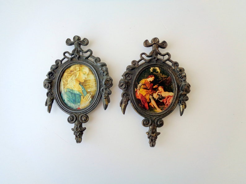 May include: Two antique oval wall plaques with ornate, dark metal frames. Each plaque features a classical painting. One depicts a woman in prayer, the other a scene with figures in a landscape. Decorative wall art.
