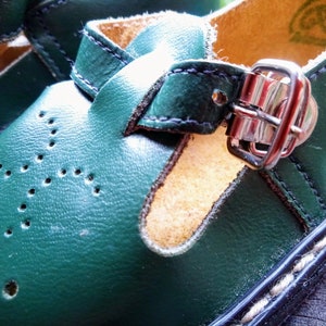 Pode incluir: Um par de sapatos de couro verde com fecho de fivela. Os sapatos t&ecirc;m um detalhe de costura branca ao redor da borda da sola.