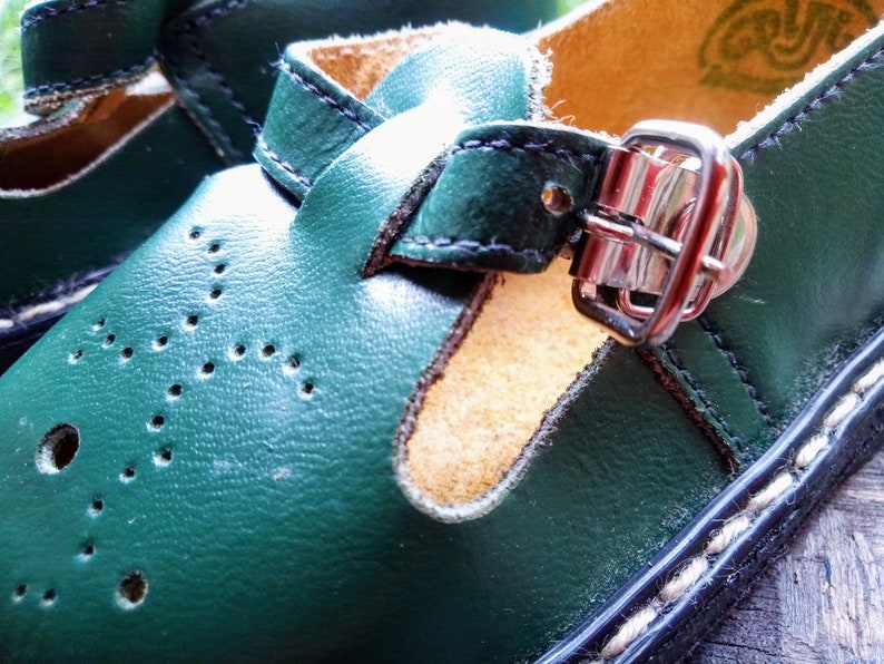Pode incluir: Um par de sapatos de couro verde com fecho de fivela. Os sapatos t&ecirc;m um detalhe de costura branca ao redor da borda da sola.