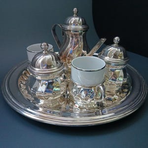 May include: A silver-toned tea set consisting of a lidded teapot, two lidded sugar bowls, and two white ceramic cups with silver rims, all arranged on a round silver-toned serving tray. The metal pieces feature a reflective, polished finish.