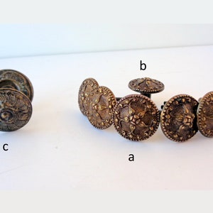 May include: A set of six antique brass buttons with intricate floral designs. The buttons are arranged in a circle, with one button in the centre. The buttons are all the same size and shape, and they have a slightly raised design.