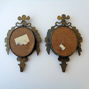May include: Two antique brass-toned wall sconces with oval frames and ornate floral and scroll designs. Each sconce has a circular opening with a cork backing and a small piece of tape. The sconces are displayed against a white background.