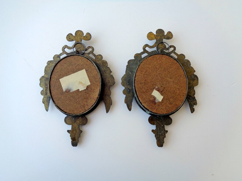 May include: Two antique brass-toned wall sconces with oval frames and ornate floral and scroll designs. Each sconce has a circular opening with a cork backing and a small piece of tape. The sconces are displayed against a white background.