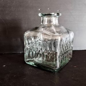 May include: A clear glass decanter with a square body and a textured surface. The decanter has a wide mouth and a short neck.