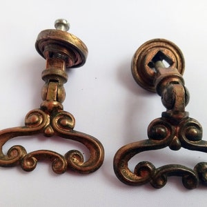 May include: Two antique brass cabinet door pulls with ornate scrollwork and a round base. The pulls have a swiveling design and are attached with screws.