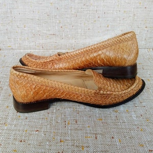 May include: A pair of tan leather loafers with a snakeskin pattern. The shoes have a low heel and a classic slip-on design. The leather has a textured appearance, and the soles are dark brown.