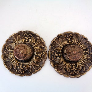 May include: Two ornate gold-toned metal decorative accents with intricate floral designs. Each accent features a raised center piece with a smaller floral design.