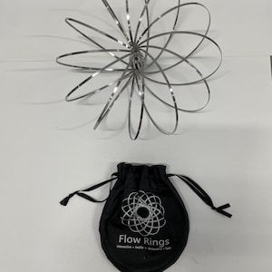 Peut inclure: Un jouet Flow Ring argenté en forme de fleur, avec un sac en tissu noir en dessous. Le sac porte l'inscription "Flow Rings" et d'autres textes. Le jouet est fait d'anneaux métalliques interconnectés.