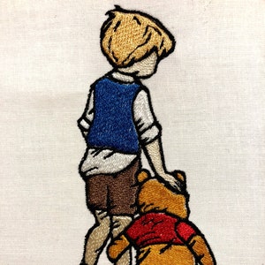 May include: Embroidered image of a boy with blond hair wearing a blue vest and brown shorts walking with a brown teddy bear wearing a red shirt.