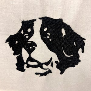 Op de afbeelding: Zwart-wit geborduurd hondenportret op crèmekleurige stof. De kenmerken van de hond zijn gedetailleerd met zwart garen, wat een textuureffect creëert. Het ontwerp is gecentreerd en toont de ogen, neus en mond van de hond.