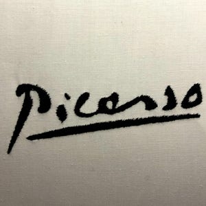 May include: Black embroidered "Picasso" signature on a white fabric. The cursive signature has a line beneath it. The fabric has a textured appearance.