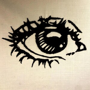 Puede incluir: Diseño de ojo bordado en negro sobre tela color crema. El ojo presenta pestañas detalladas, un iris definido y una pupila prominente. El bordado se realiza con hilo grueso y oscuro, creando un contraste audaz contra el fondo claro.