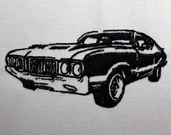 May include: Black and white embroidered image of a classic car. The car has a long bonnet and a rounded front end. The car is facing the viewer.
