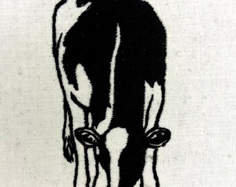 May include: Black and white embroidered image of a cow grazing, set against a white fabric background. The cow is depicted in a stylized, simplified form, with its head lowered towards the ground.