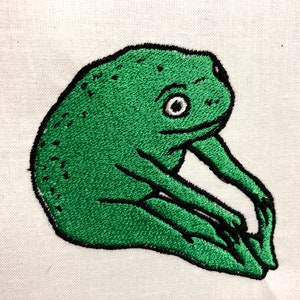 May include: Green embroidered frog with black outlines, sitting on a white fabric background.