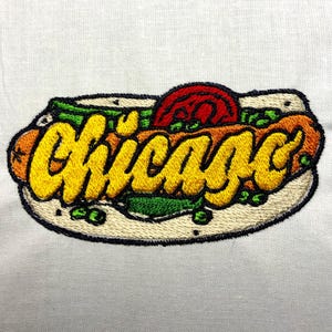 May include: Embroidered patch of a hot dog with toppings, the word "Chicago" is written in yellow cursive across the hot dog.