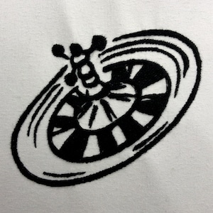 May include: Black embroidered design on white fabric. The design features a stylized wheel with radiating lines and a cluster of small circles. The wheel has alternating black and white segments, creating a bold contrast.