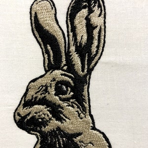 May include: Embroidered image of a rabbit's head and ears, featuring detailed stitching in black and tan against a white background. The rabbit's profile is visible, showcasing its eye and long ears. The design is likely for home decor or textile applications.