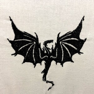 May include: Black embroidered silhouette of a dragon with wings spread wide, on a white fabric background.