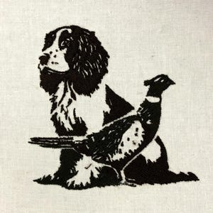Puede incluir: Diseño bordado en blanco y negro de un perro y un faisán sobre tela color crema. El perro es un spaniel, el faisán está detallado con manchas y plumas. El bordado se realiza con hilo grueso y oscuro, creando un efecto texturizado.
