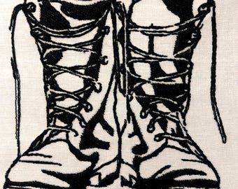 Op de afbeelding: Zwarte geborduurde illustratie van twee gevechtslaarzen met veters op een witte achtergrond. De laarzen zijn gedetailleerd met stiksels, die de vorm en textuur van het schoeisel laten zien. De veters zijn lang en hangen naar beneden.