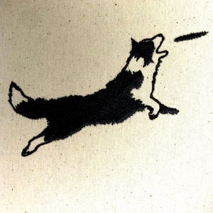 May include: Embroidered image of a black and white dog leaping to catch a frisbee. The dog is in mid-air, mouth open, with the frisbee in front. The embroidery is on a beige fabric.