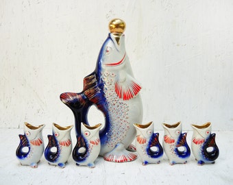May include: A set of seven porcelain fish-shaped shot glasses. The largest fish is a decanter with a gold top. The smaller fish are painted in shades of blue, white, and red.