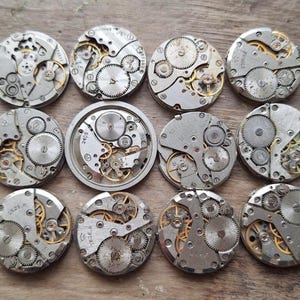 May include: A collection of twelve vintage watch movements, showcasing intricate gears, springs, and other mechanical components. The movements are primarily silver-toned with gold accents, arranged on a wooden surface. The gears are visible, highlighting the craftsmanship.