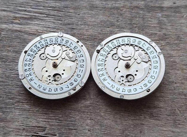 K&ouml;nnte beinhalten: Zwei runde Vintage-Uhrwerkkomponenten mit komplexen Zahnr&auml;dern und Datumsanzeigen. Die silberfarbenen Teile weisen kleine Schrauben und Zahlen von 1 bis 31 auf, die die Tage des Monats anzeigen. Auf einem Holzgrund.