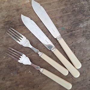 May include: A set of antique silverware, including two forks and two knives, arranged on a wooden surface. The knives have decorative blades and cream-colored handles. The forks and knives are made of silver-colored metal.