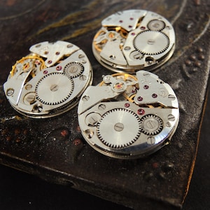Pode incluir: Tr&ecirc;s movimentos de rel&oacute;gio vintage com engrenagens intrincadas, componentes de metal prateado e pequenas joias vermelhas. As pe&ccedil;as do rel&oacute;gio est&atilde;o dispostas sobre uma superf&iacute;cie escura e texturizada, mostrando a mec&acirc;nica detalhada dos rel&oacute;gios.