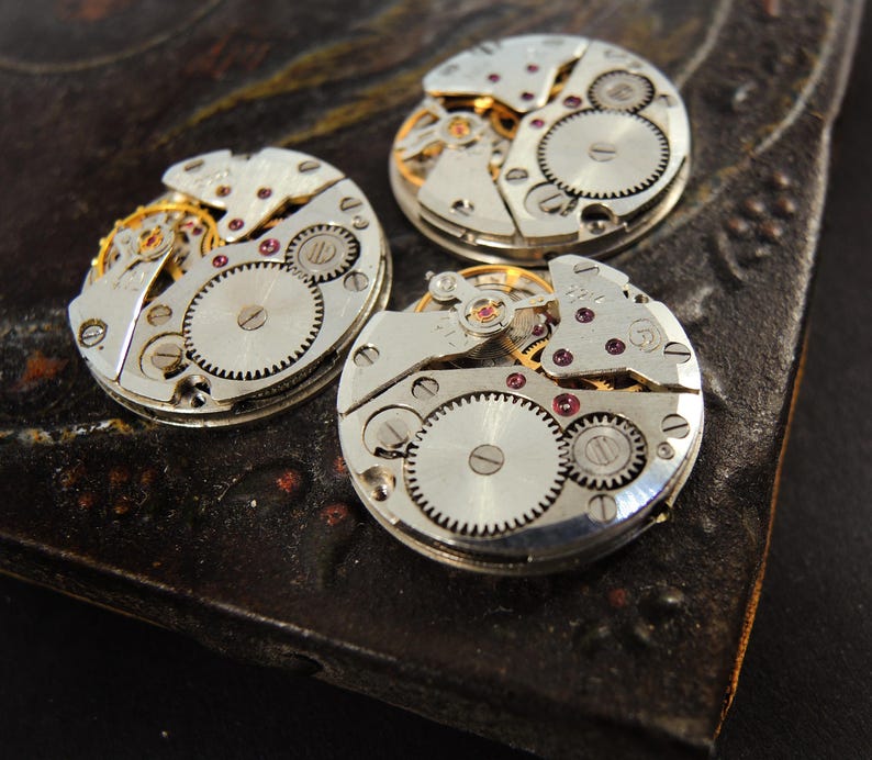 Pode incluir: Tr&ecirc;s movimentos de rel&oacute;gio vintage com engrenagens intrincadas, componentes de metal prateado e pequenas joias vermelhas. As pe&ccedil;as do rel&oacute;gio est&atilde;o dispostas sobre uma superf&iacute;cie escura e texturizada, mostrando a mec&acirc;nica detalhada dos rel&oacute;gios.