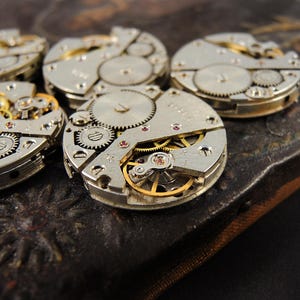 May include: Close-up of several disassembled watch movements, showcasing intricate gears, springs, and jewels. The silver-toned components are arranged on a dark, textured surface, highlighting the mechanical details of the timepieces.
