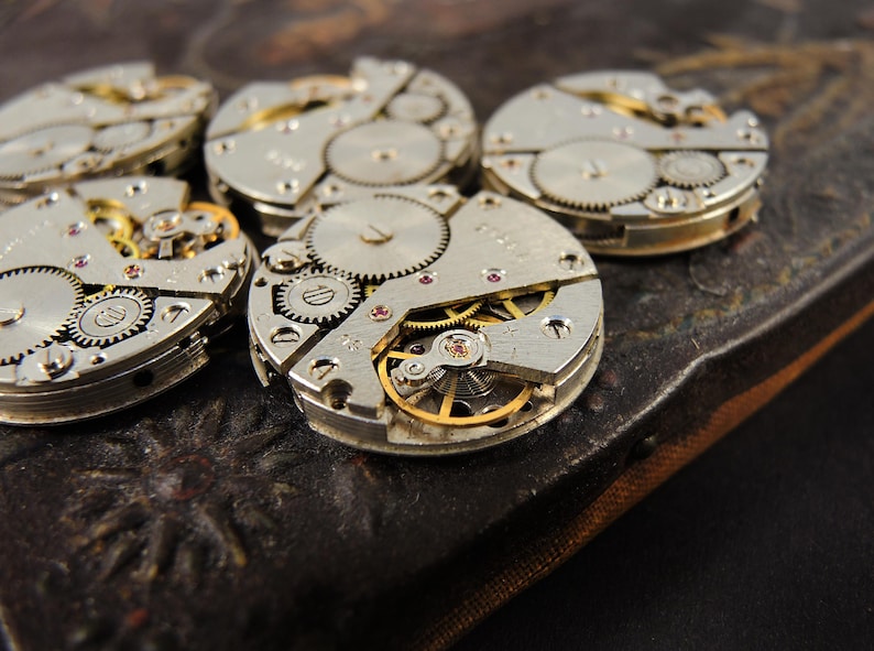 May include: Close-up of several disassembled watch movements, showcasing intricate gears, springs, and jewels. The silver-toned components are arranged on a dark, textured surface, highlighting the mechanical details of the timepieces.