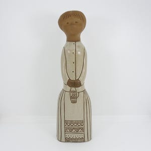 May include: A tall ceramic figurine of a person with a smooth brown head and hair. The body is a light beige color with dark brown line drawings depicting a buttoned shirt, hands clasped in front, and a skirt with geometric patterns at the hem.