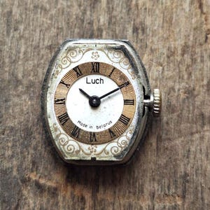 May include: Vintage Luch wristwatch with a silver-tone case and a white and gold face. The watch features black Roman numerals and hands, with the text "Luch" and "Made in Belarus" visible. The watch has a textured crown.