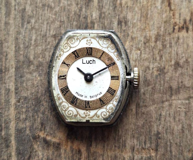 May include: Vintage Luch wristwatch with a silver-tone case and a white and gold face. The watch features black Roman numerals and hands, with the text "Luch" and "Made in Belarus" visible. The watch has a textured crown.
