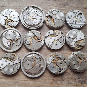 May include: A collection of twelve disassembled watch movements, showcasing intricate gears and mechanisms. The silver-toned components are arranged on a wooden surface, with visible brass-colored gears adding contrast. The circular shapes and detailed inner workings are prominent.