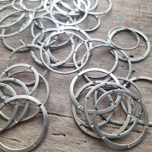 May include: A collection of silver-colored metal rings with a circular design. The rings are scattered across a wooden surface, creating a textured and layered effect. The rings have a cut-out design.